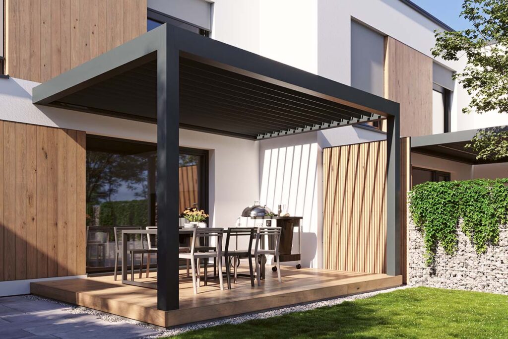 Moderní antracitová bioklimatická pergola zastřešuje venkovní jídelnu s grilem na dřevěné terase u domu s dřevěným obkladem fasády — srovnání dřevěná nebo hliníková pergola v moderním kontextu.