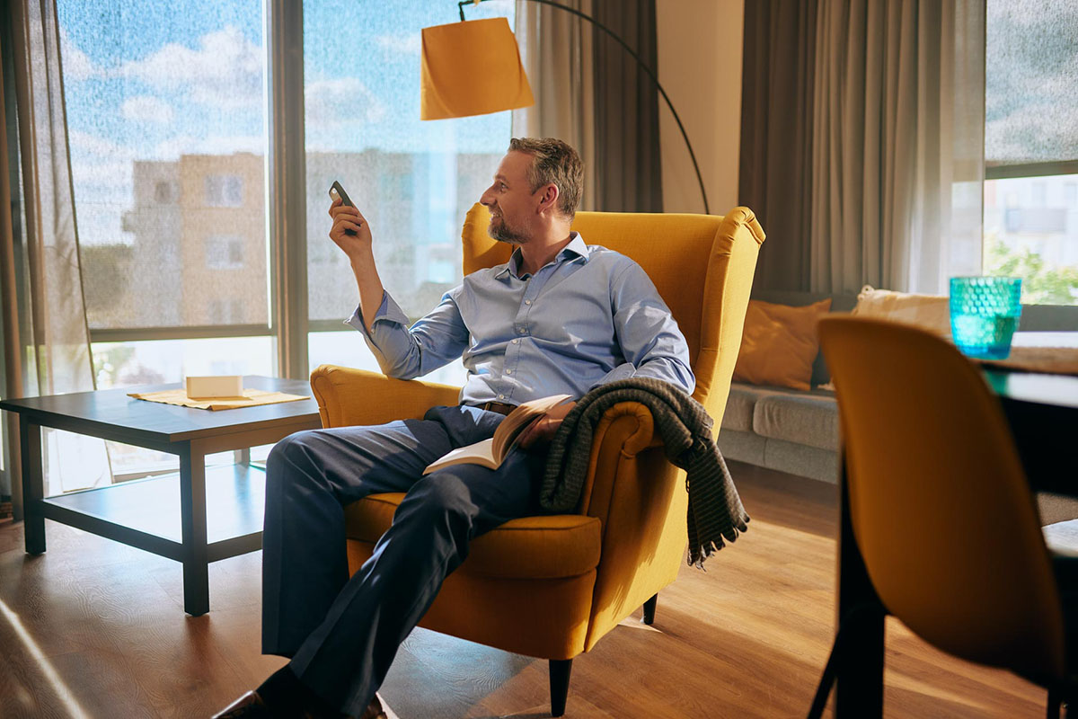 Screenové rolety v pozadí, obývají pokoj a sedící muž, který dálkovým ovladačem ovládá stínění