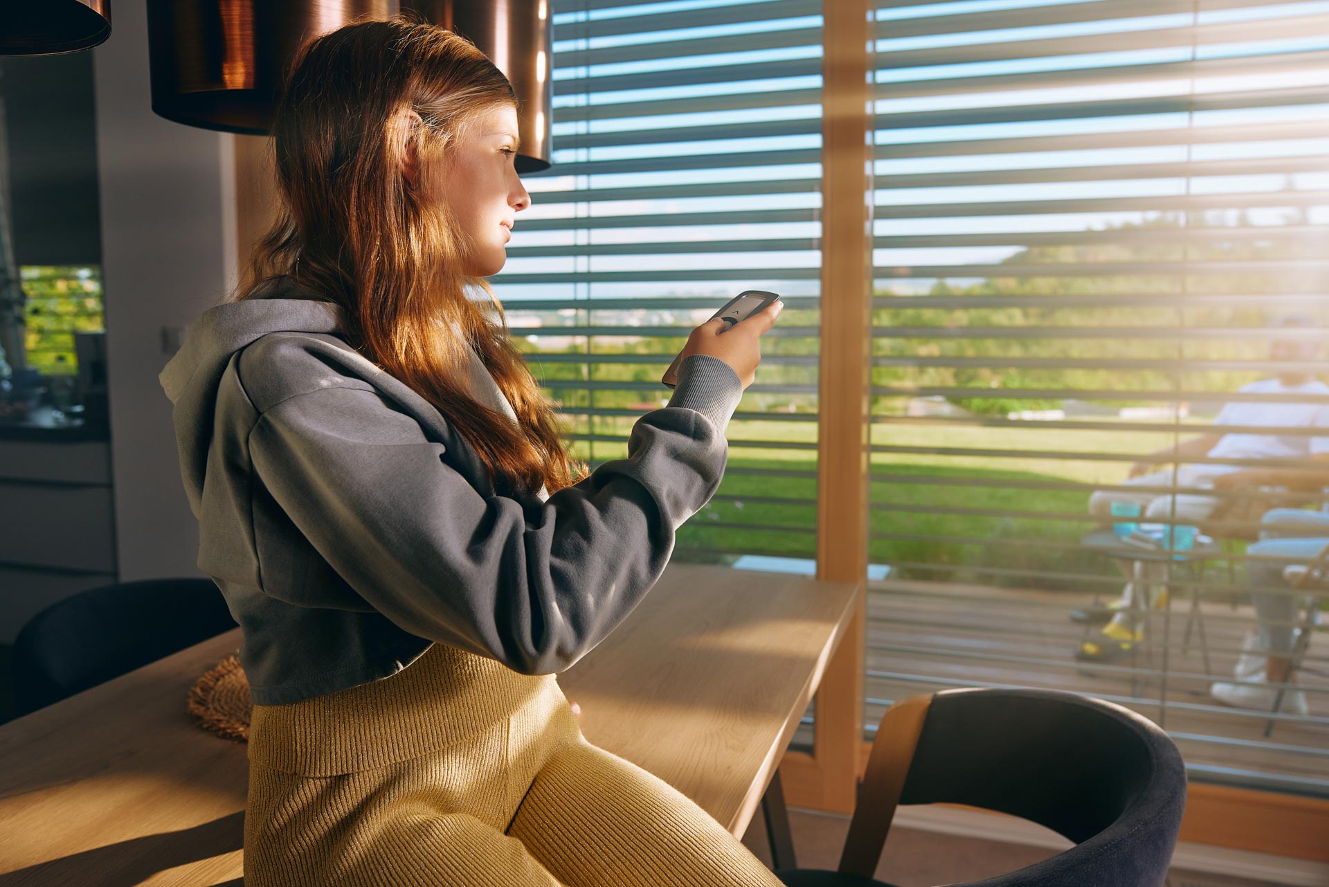 Žena sedí na kuchyňském pultu a ovladačem nastavuje venkovní žaluzie